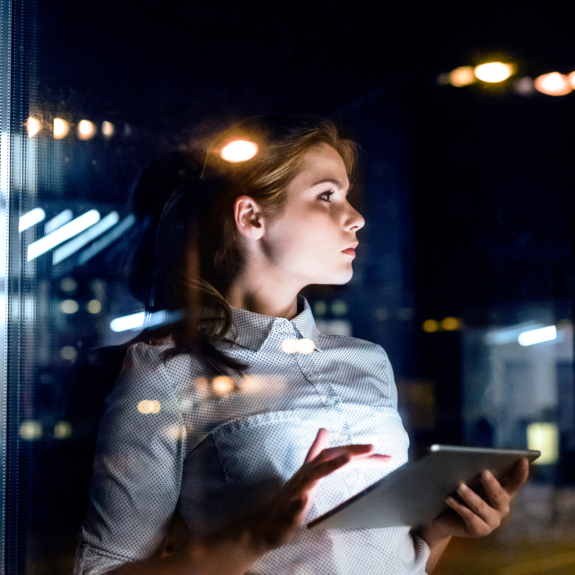 young woman entrepreneur holding a tablet seen through office window during the evening