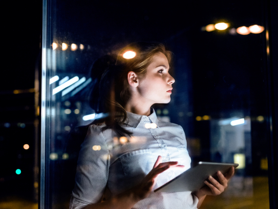 young woman entrepreneur holding a tablet seen through office window during the evening